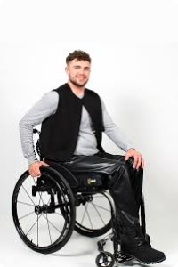 A young man in a wheelchair wearing IZ adaptive seamless back faux leather trousers, black waistcoat and long sleeved grey top.